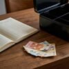 Escena de escritorio con una caja de caudales negra abierta, billetes de euro, monedas y un libro contable para ahorros domésticos.