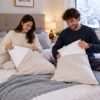 Pareja sonriente arreglando cojines decorativos sobre la cama de su dormitorio.